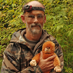 Author Philippe Coudray poses in a camo sweater and a headlamp as he holds a stuffed orange monkey